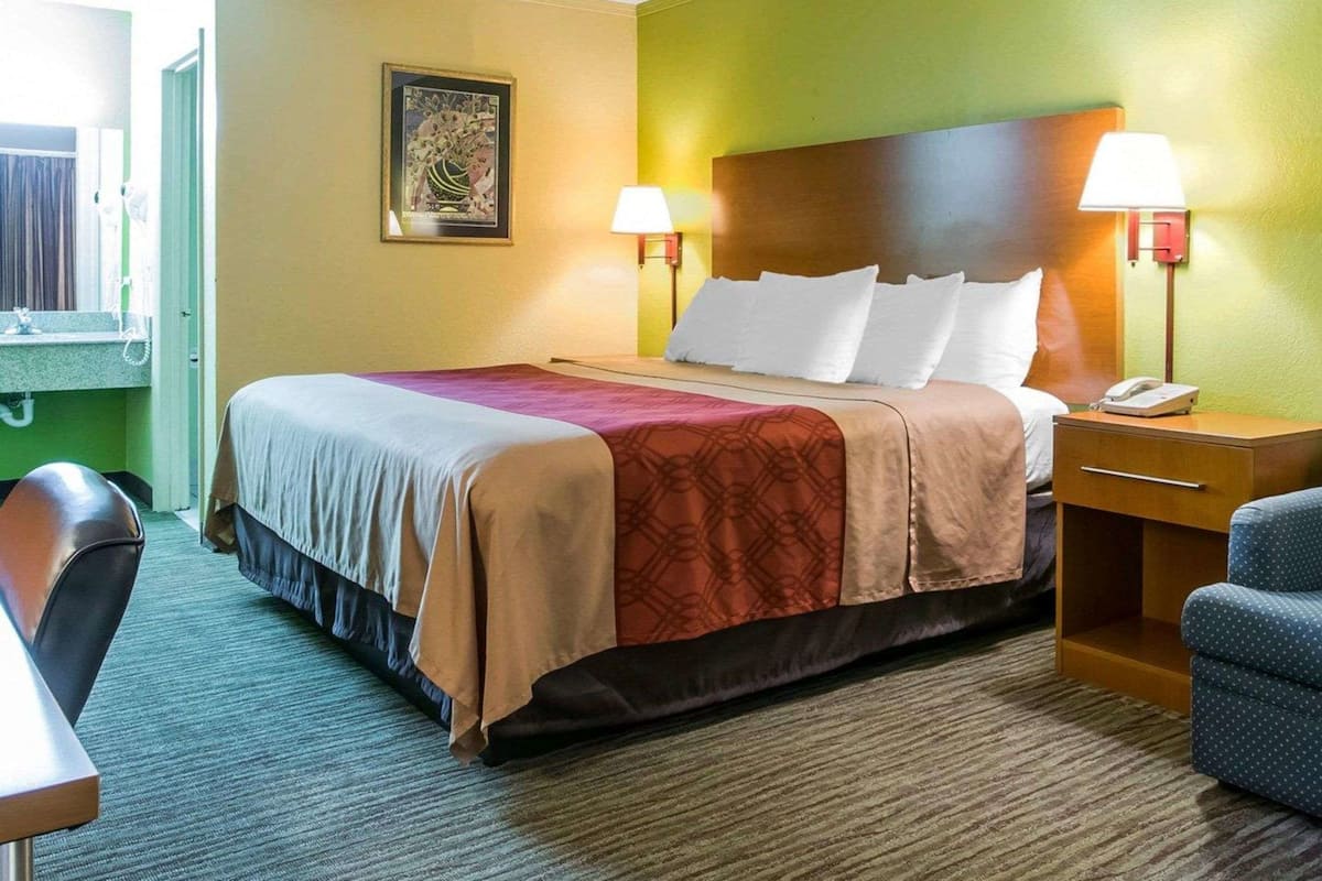 A hotel room with a bed, two bedside lamps, a nightstand with a phone, a chair, and a painting on the wall.
