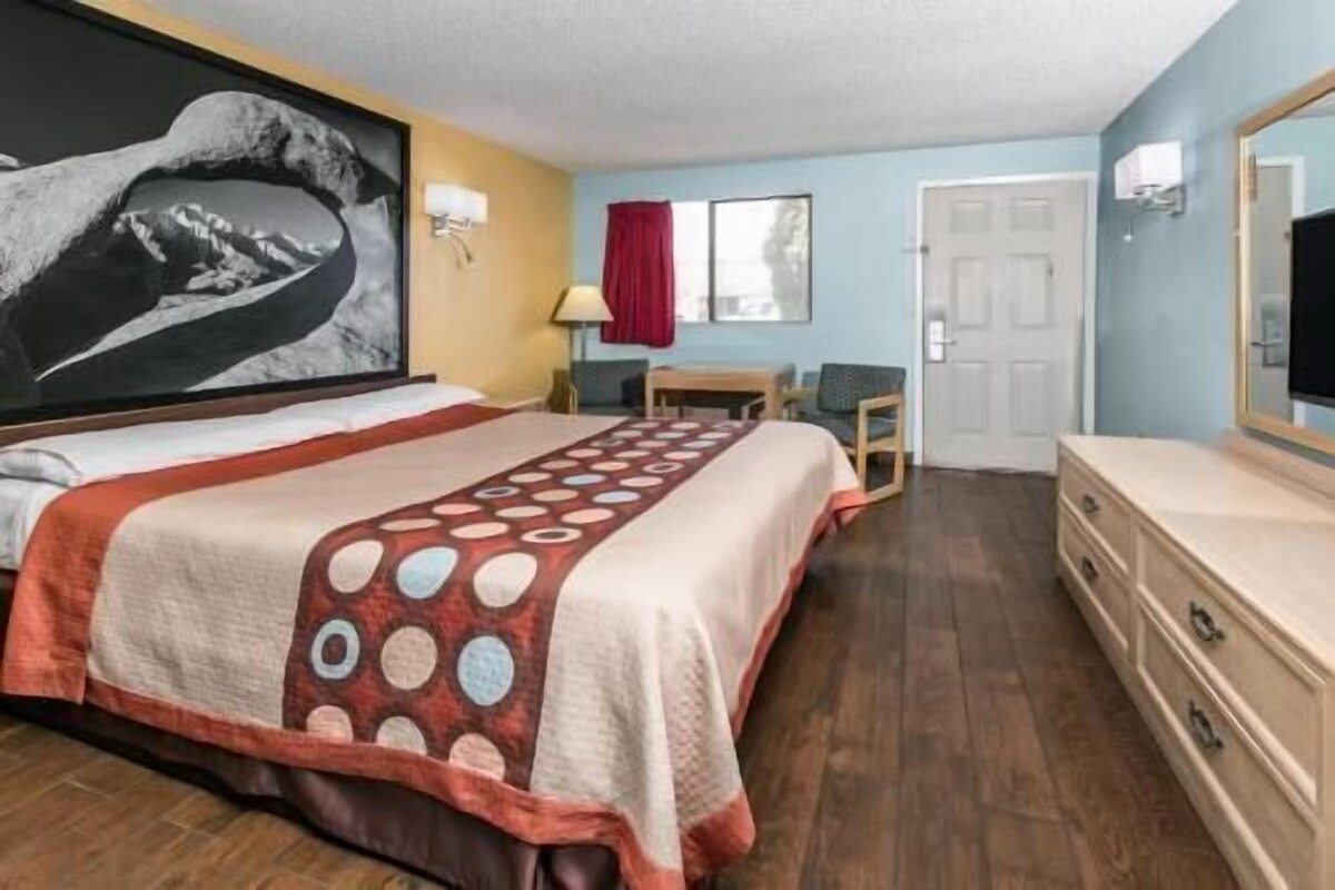 Habitación de hotel con una cama, un aparador, un escritorio, una silla, un ventanal con cortina roja y un televisor de pared.