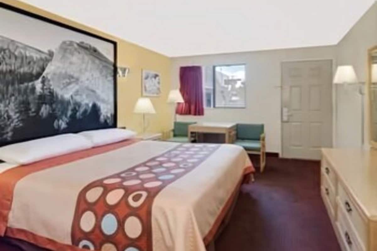 A hotel room with a bed, a desk, a chair, a lamp, a painting, and a window.