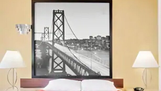 Habitación de hotel con una cama, dos lámparas de noche, un cuadro de un puente y una mesita de noche con lámpara.