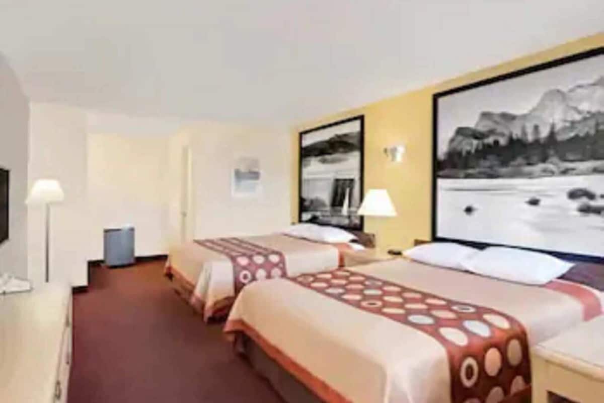 A hotel room with two beds, a desk, a chair, a lamp, and a painting of a mountain landscape.