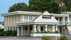 Ein zweigeschossiges Motel mit einem Schild, auf dem "RELAXNN Cape Code Inn" steht. Es verfügt über eine überdachte Veranda und einen eingezäunten Parkplatz.