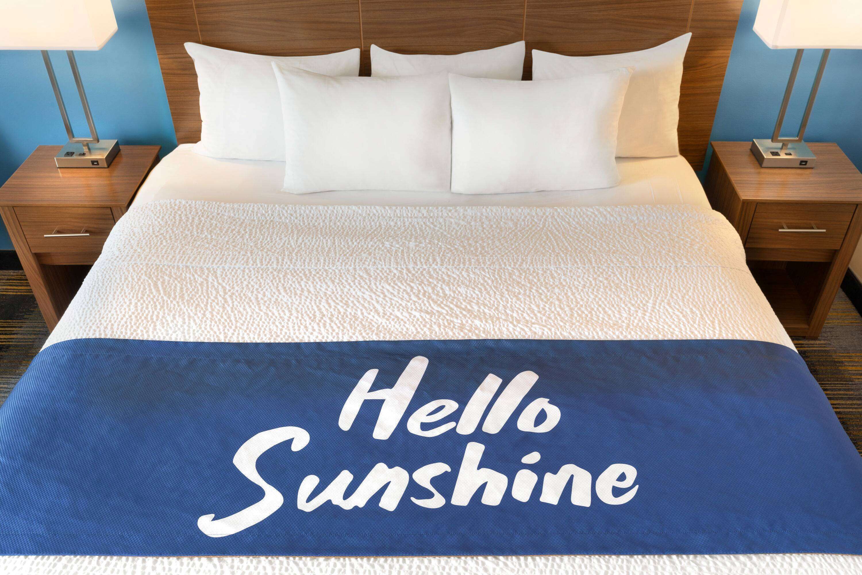 A neatly made bed with a blue blanket that reads "Hello Sunshine."