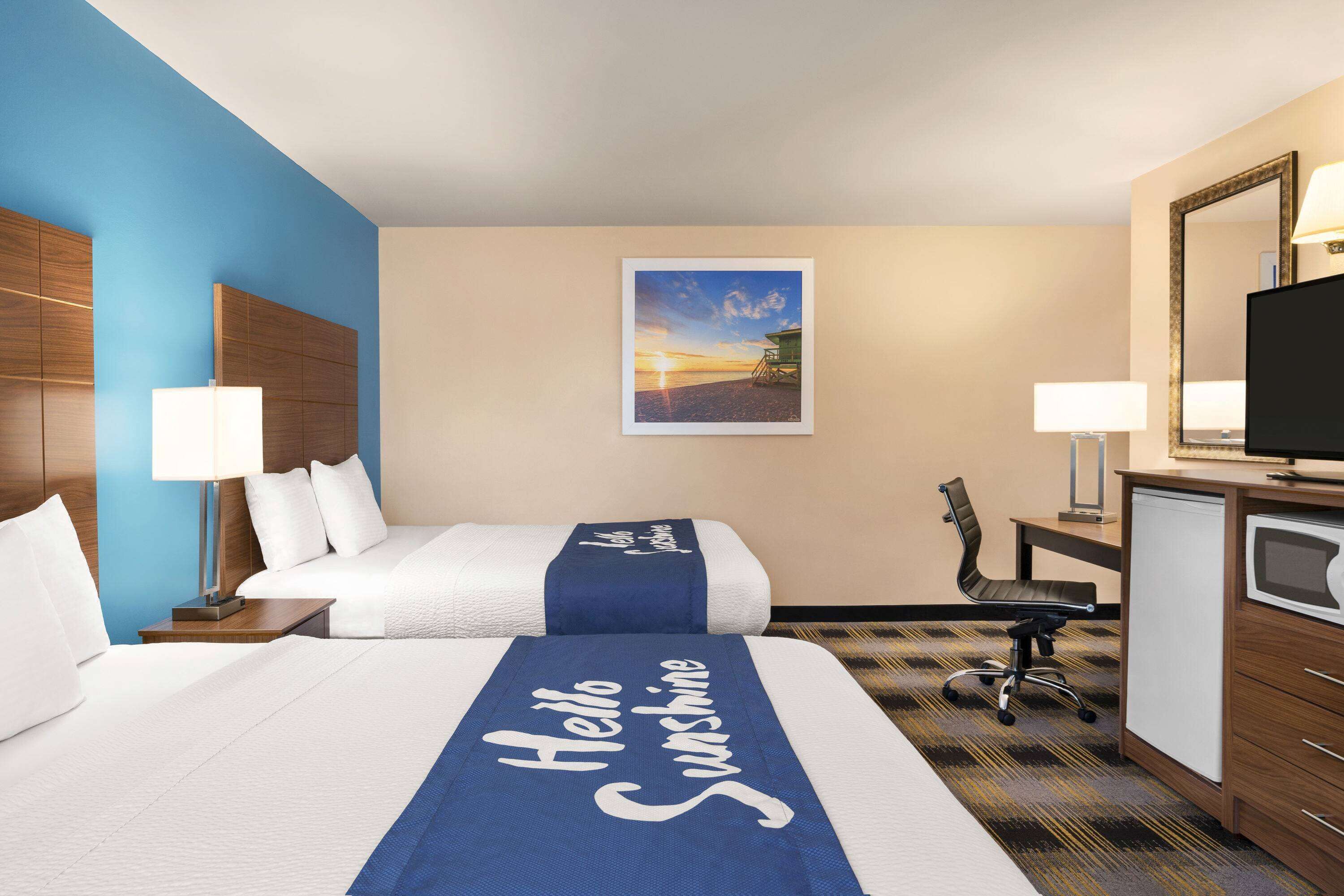 A hotel room with a bed, two bedside tables, a desk with a chair, a television, and a painting of a sunset.
