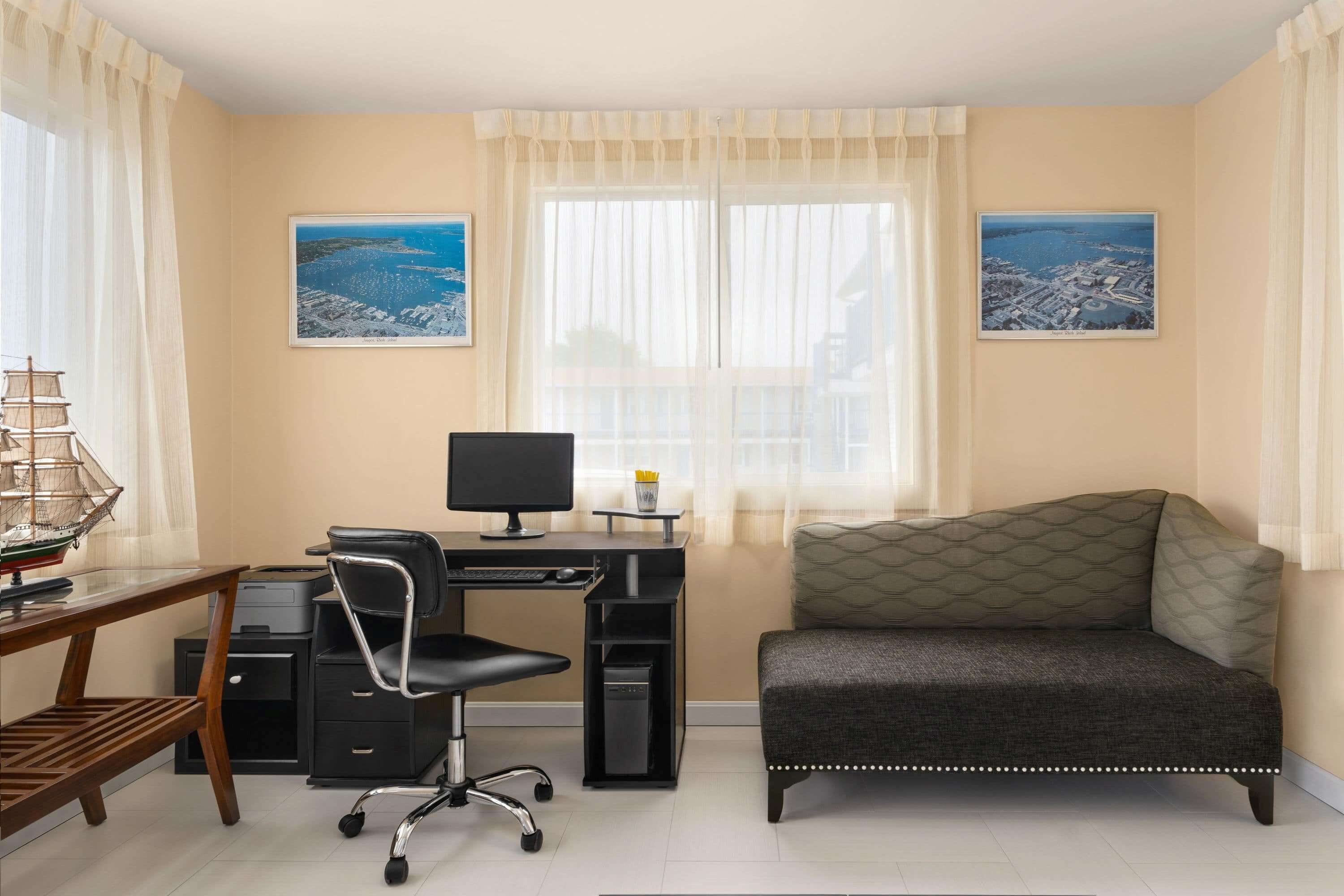 A room with a desk, computer, chair, and a sofa. There are two windows with curtains, and two framed pictures on the walls.