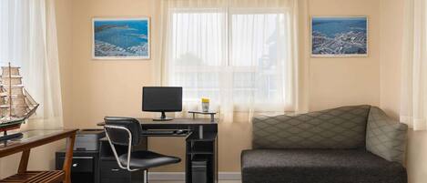 A room with a desk, computer, chair, and a sofa. There are two windows with curtains, and two framed pictures on the walls.
