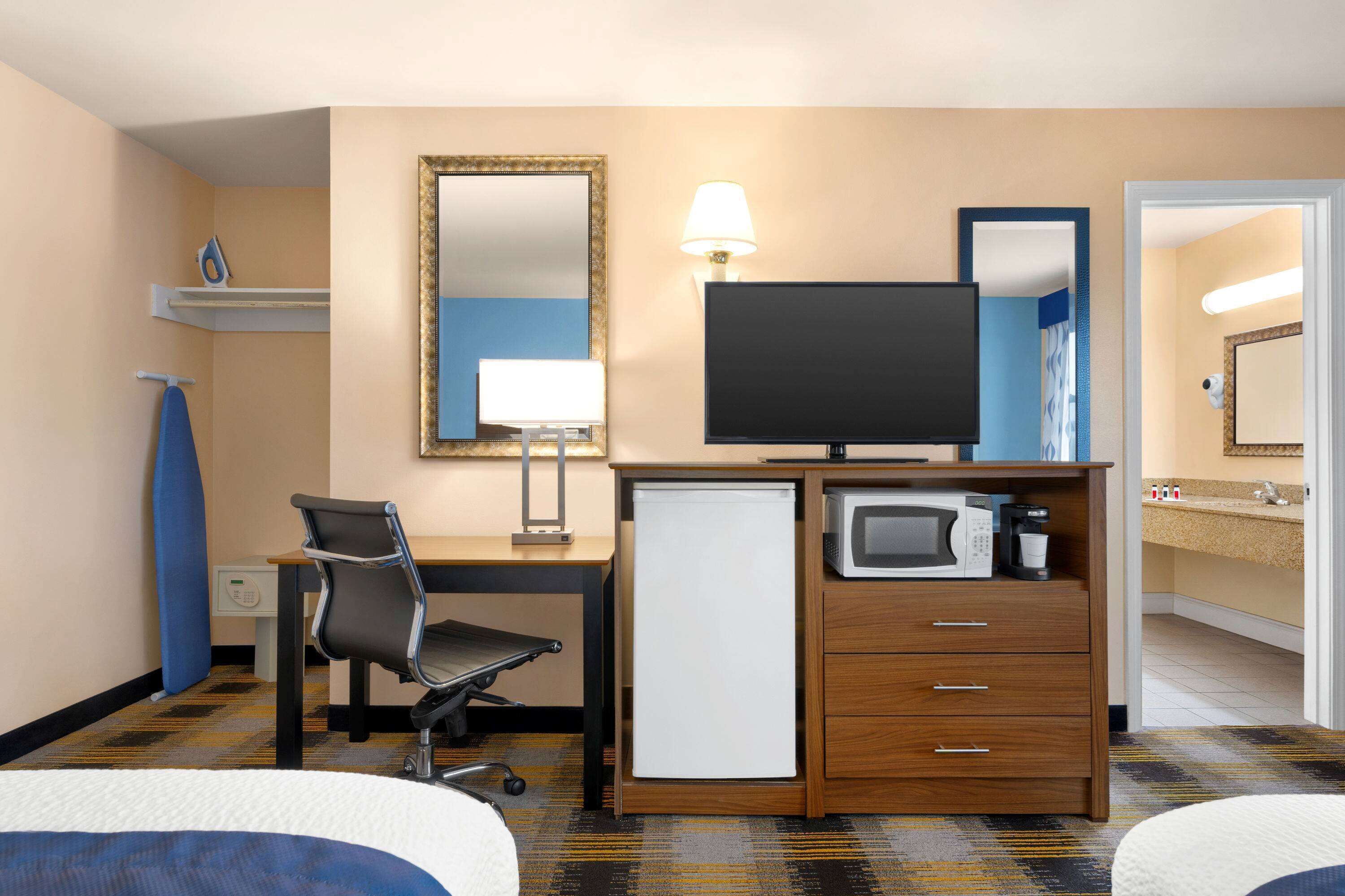 Habitación de hotel con cama, escritorio, silla, televisor, refrigerador pequeño y microondas, además de un baño con lavamanos y espejo.