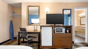 Habitación de hotel con cama, escritorio, silla, televisor, refrigerador pequeño y microondas, además de un baño con lavamanos y espejo.