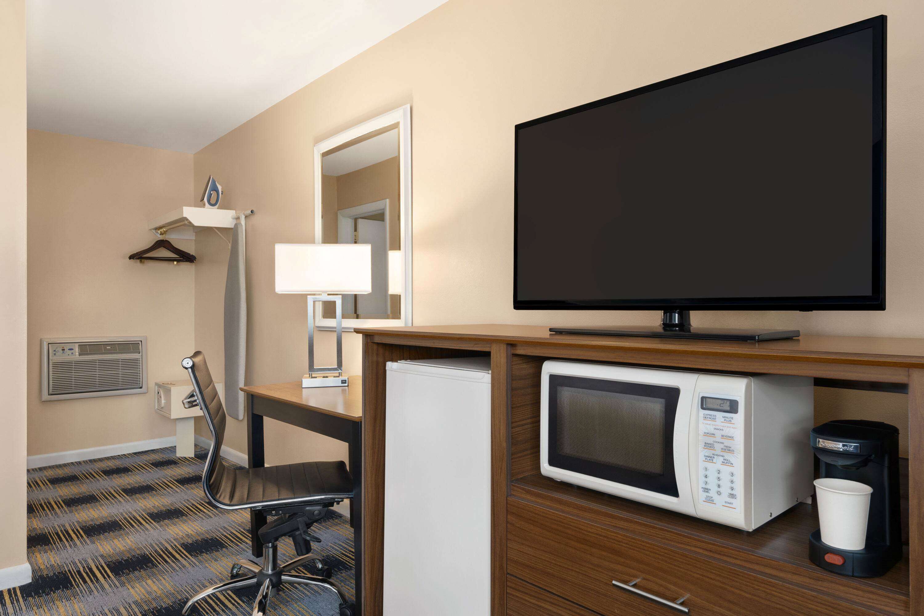 A hotel room with a flat-screen TV, a microwave, a coffee maker, a desk with a chair, a hanging coat rack, and a mirror.