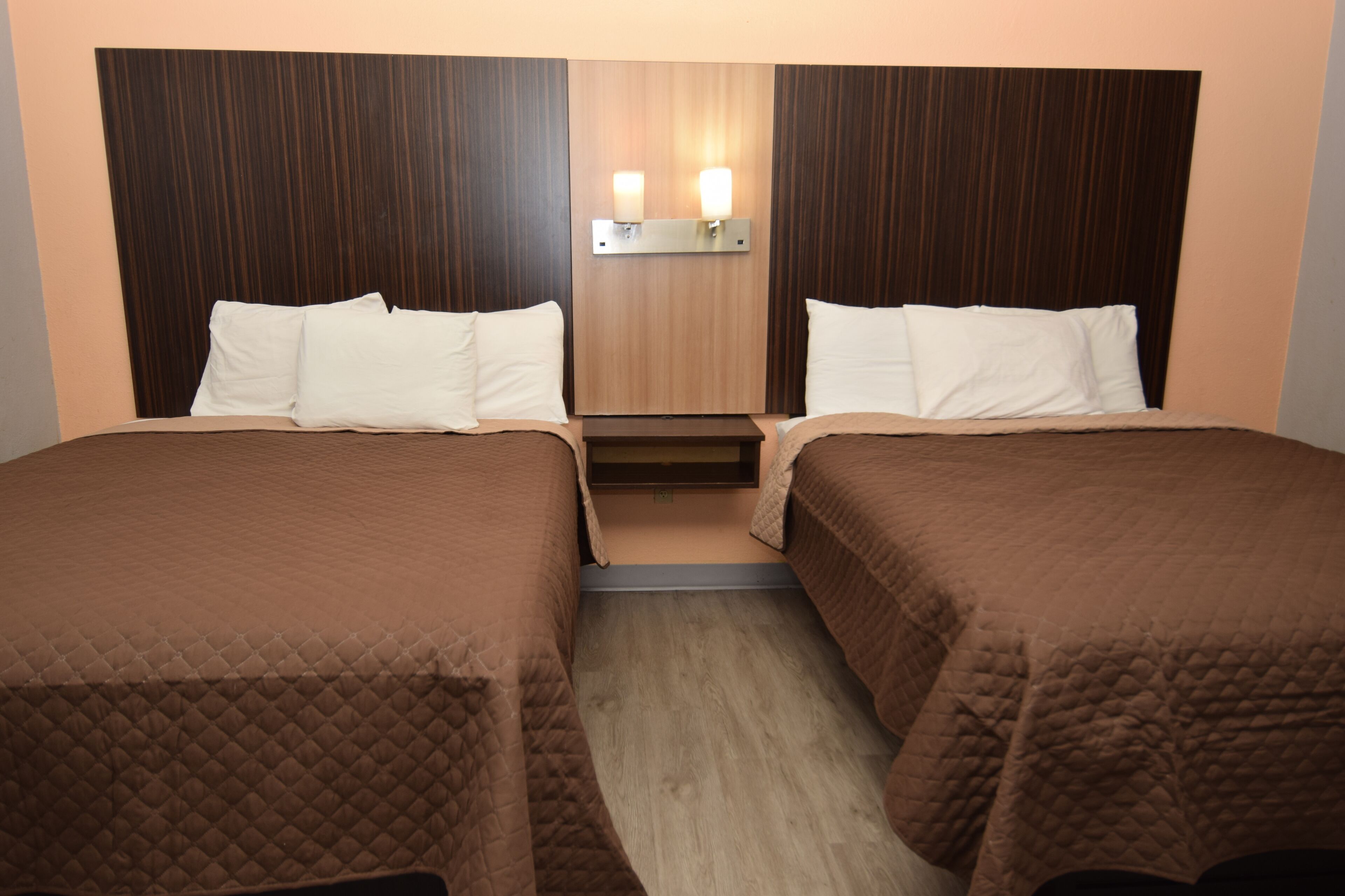 A hotel room with two beds, each with a brown quilted bedspread and white pillows. There is a wooden headboard with a light fixture above the beds.