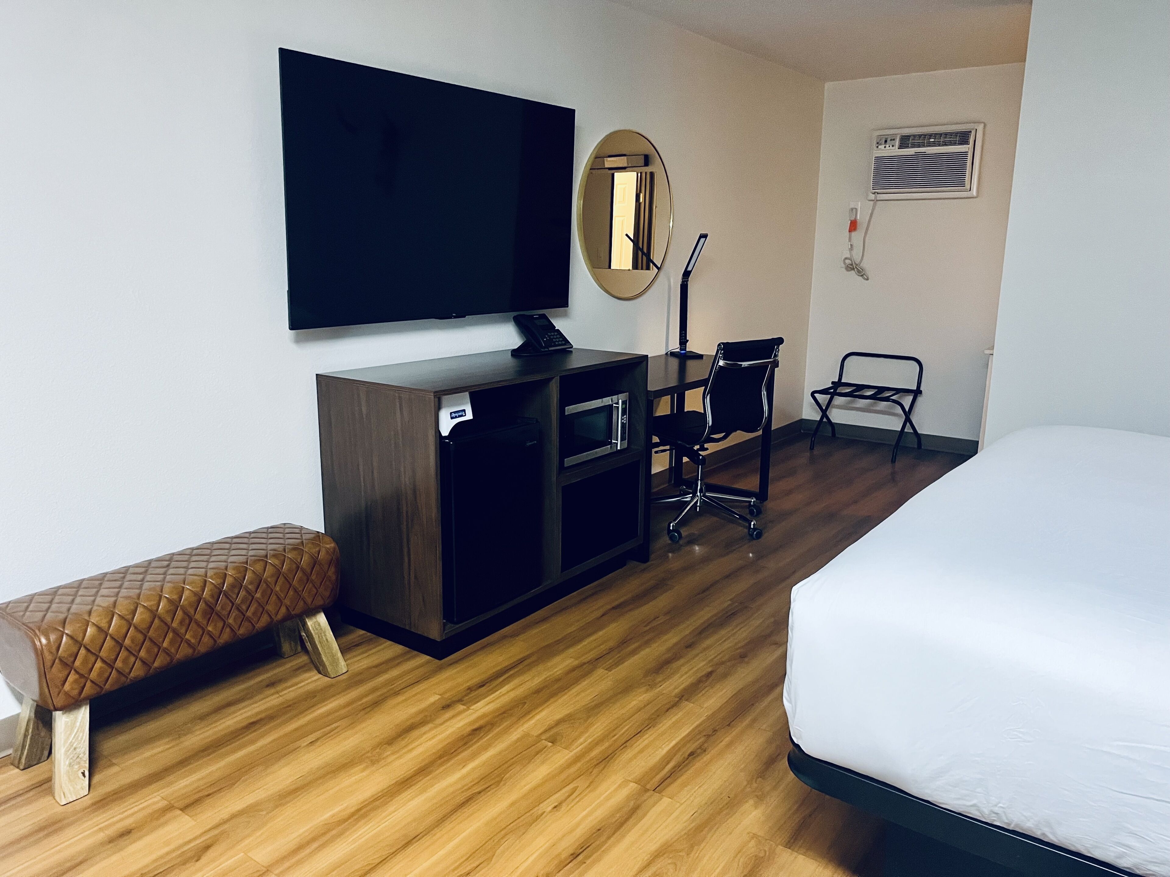 A hotel room with a bed, a desk with a chair, a television, a mirror, and a small kitchenette.