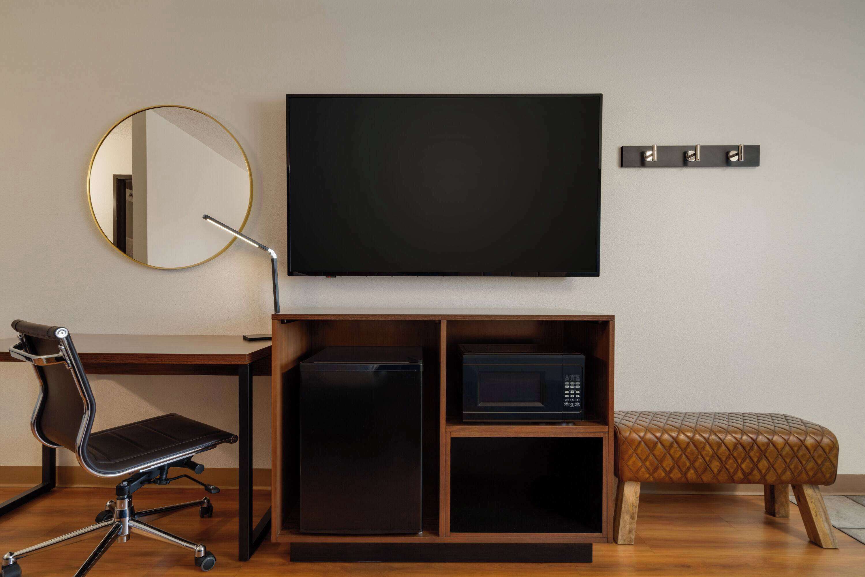 A compact room with a flat-screen TV, a small refrigerator, a microwave, a round mirror, and a cushioned bench.