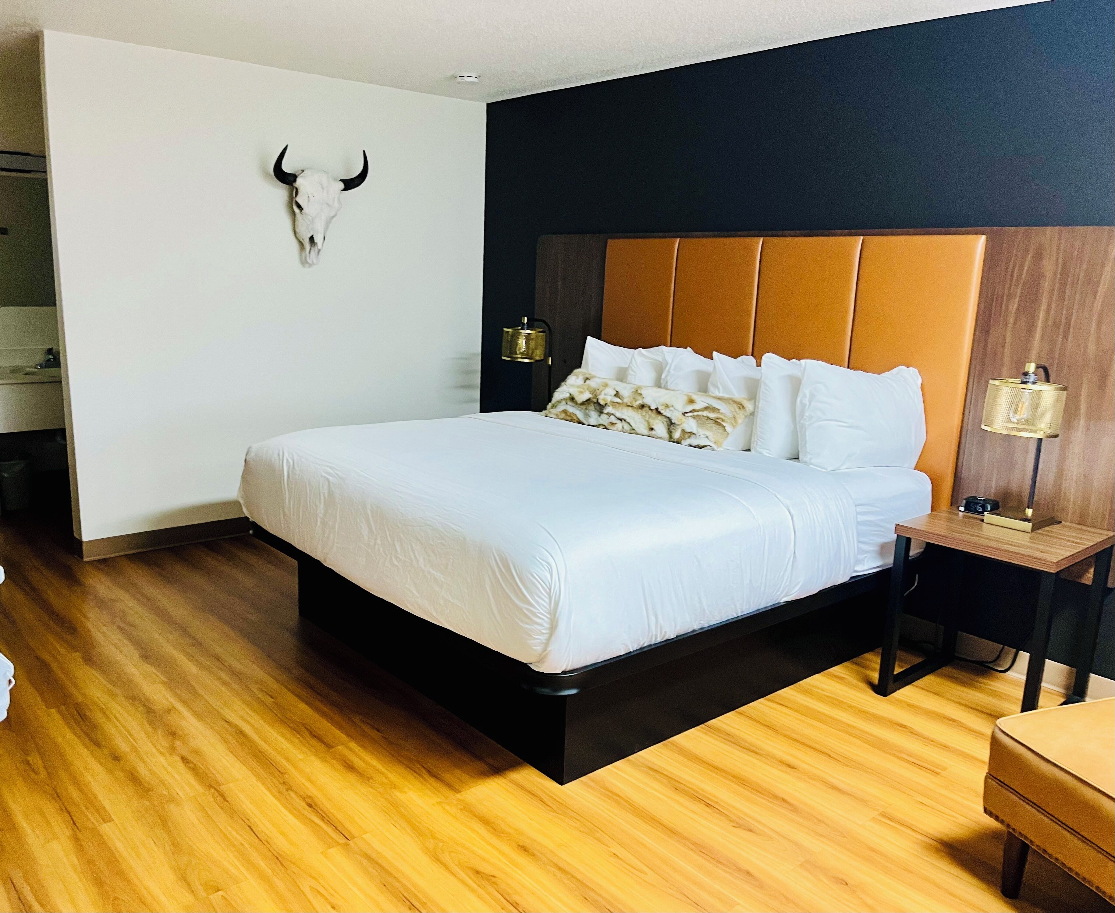 A modern hotel room with a large bed, wooden flooring, a black headboard, a bedside lamp, and a wall-mounted bull skull decoration.