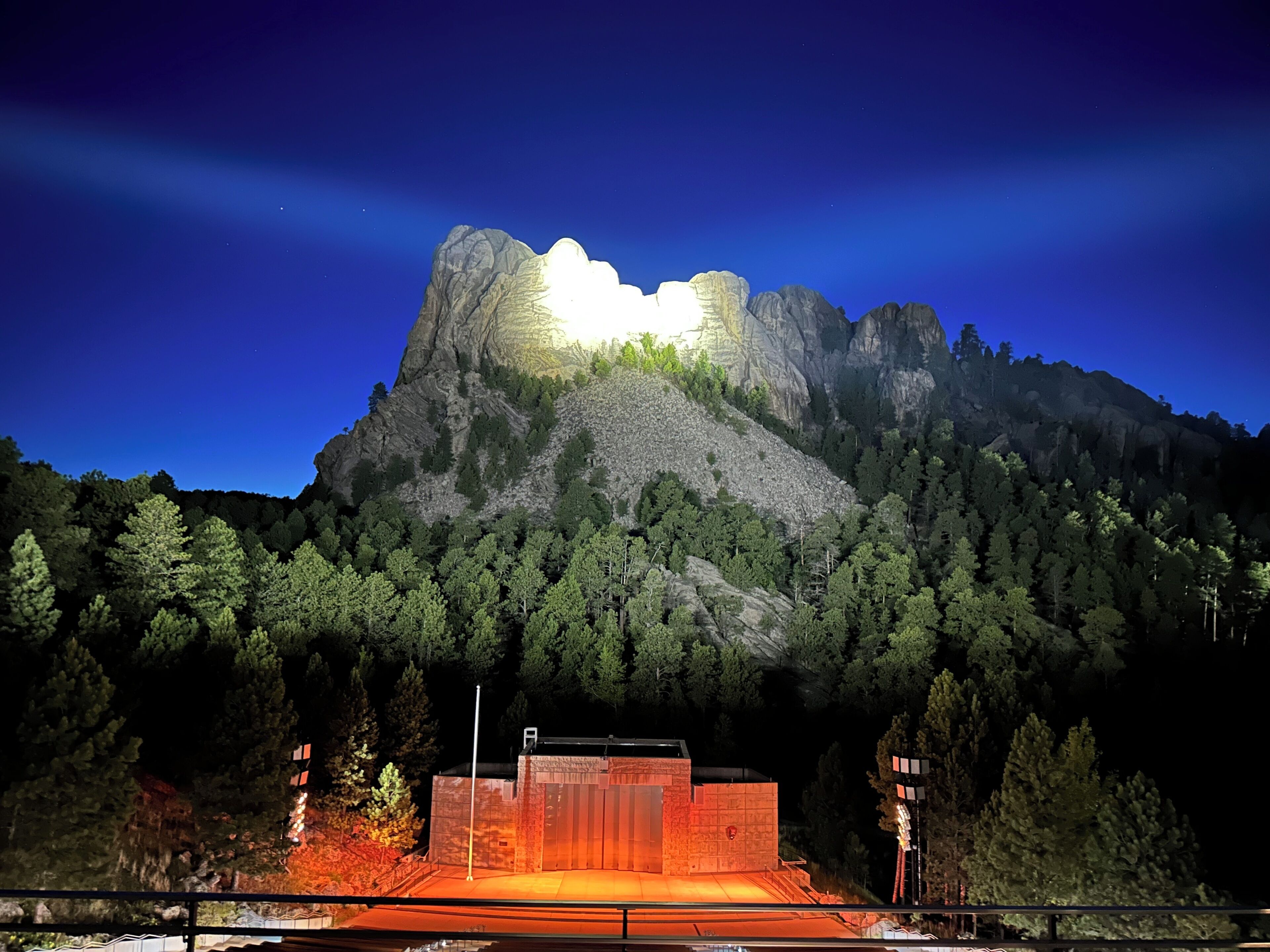 Ein Berg mit einem markanten Gipfel, ein Gebäude mit flachem Dach und ein bewaldeter Bereich im Vordergrund.