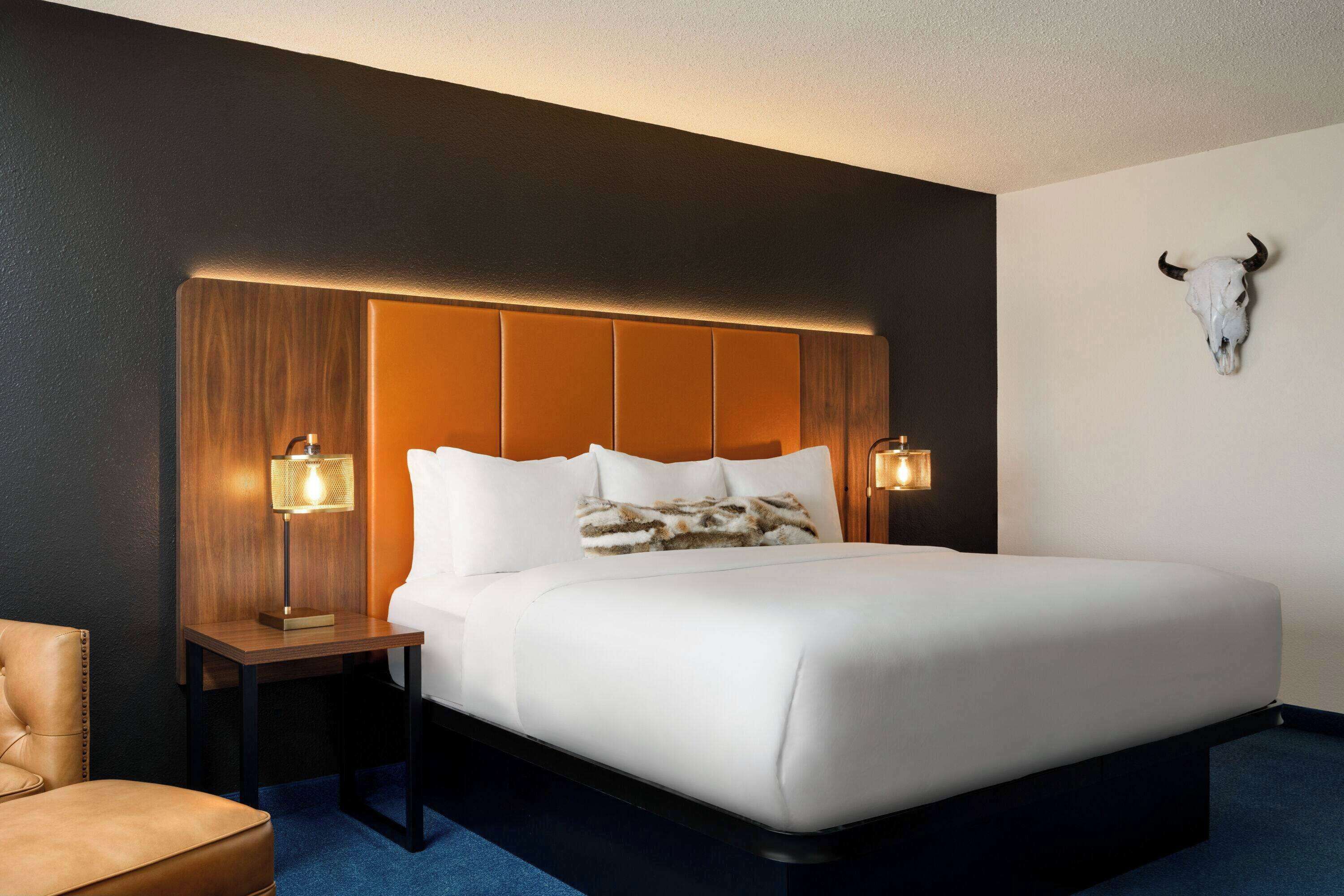 A hotel room with a large bed, two bedside tables with lamps, a wooden headboard, and a wall-mounted bull skull decoration.