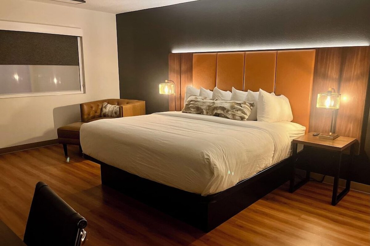 Habitación de hotel moderna con una cama grande, mesitas de noche, una silla y vistas a la ciudad.