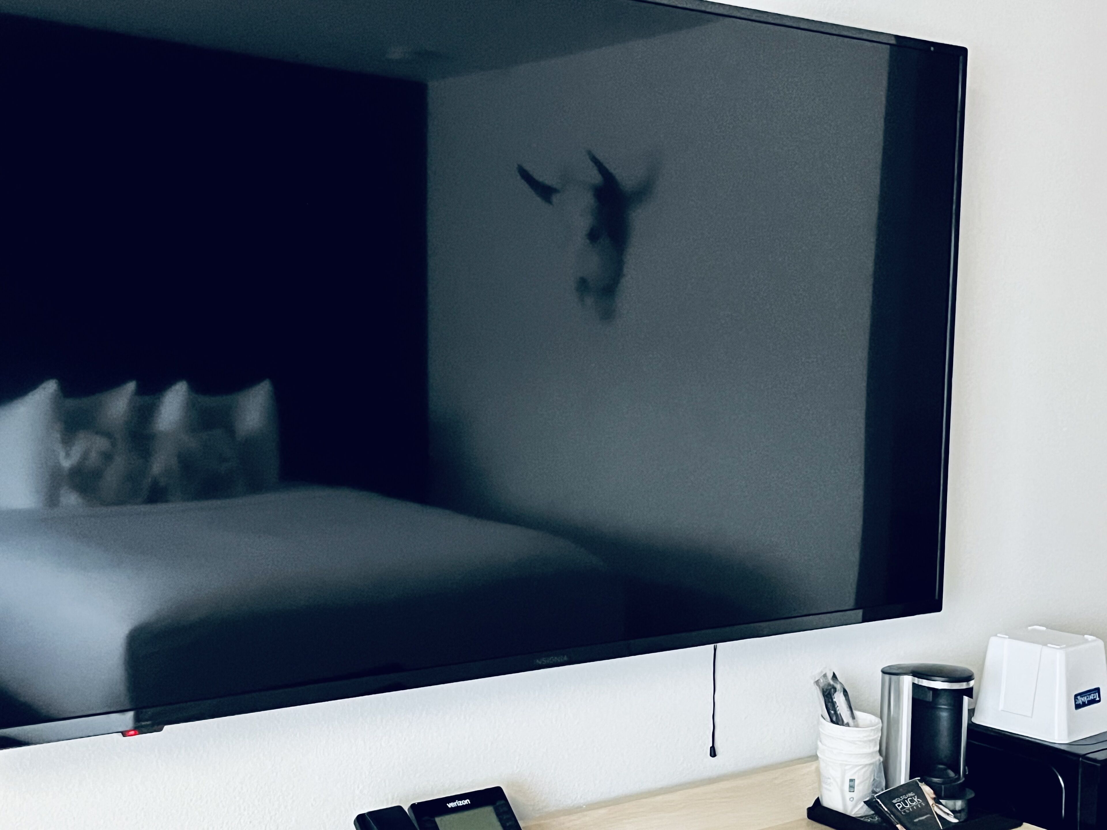 A hotel room with a large flat-screen TV mounted on the wall, a bed with pillows, a coffee maker, and a small table with a cup and a book.