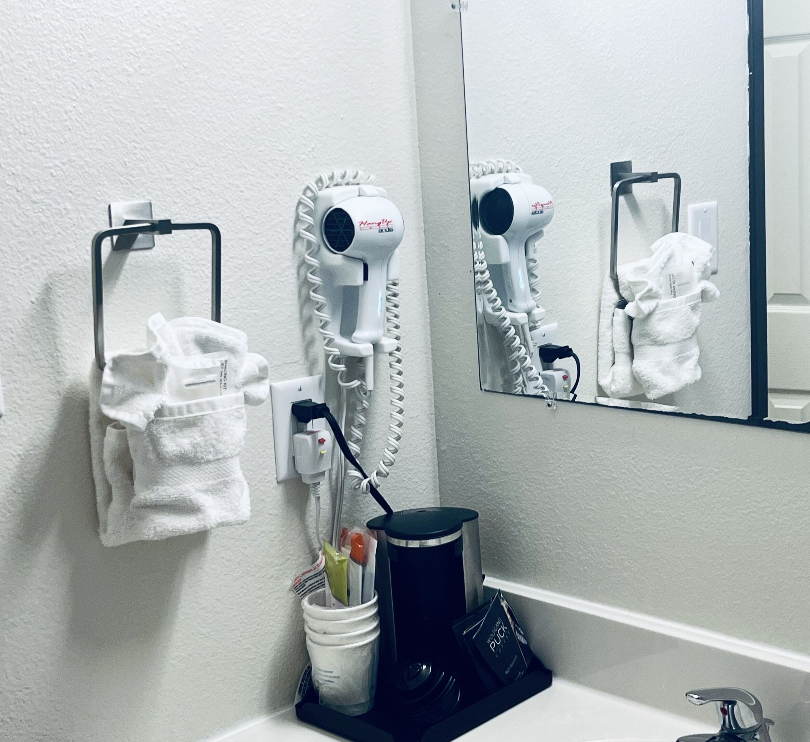 A bathroom with a towel rack, two hair dryers, a trash bin, and a coffee machine.