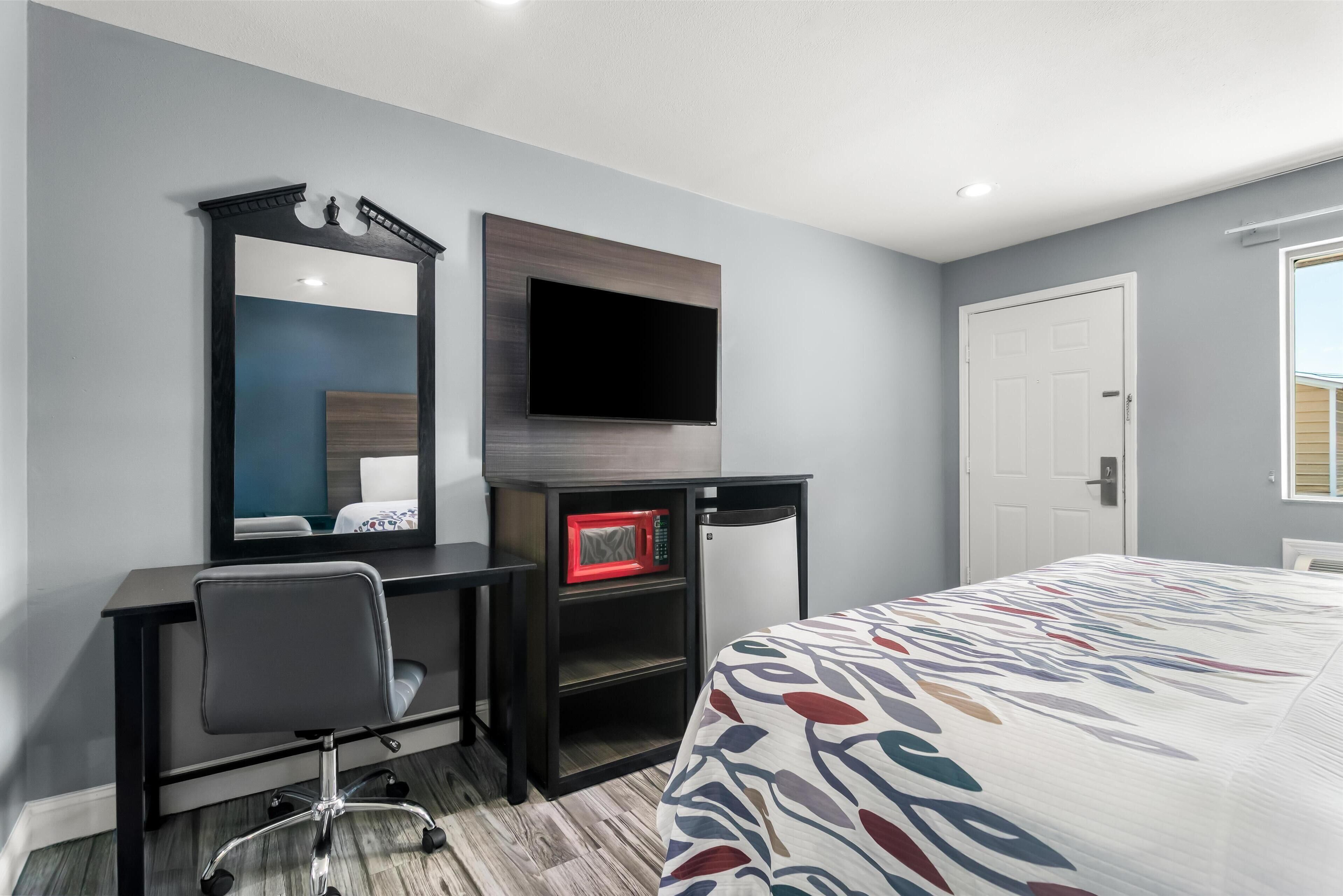 A hotel room with a bed, a desk with a chair, a television, a microwave, and a door.