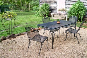 Un patio con mesa y sillas de metal, rodeado de grava, con un jardín y una construcción de madera al fondo.
