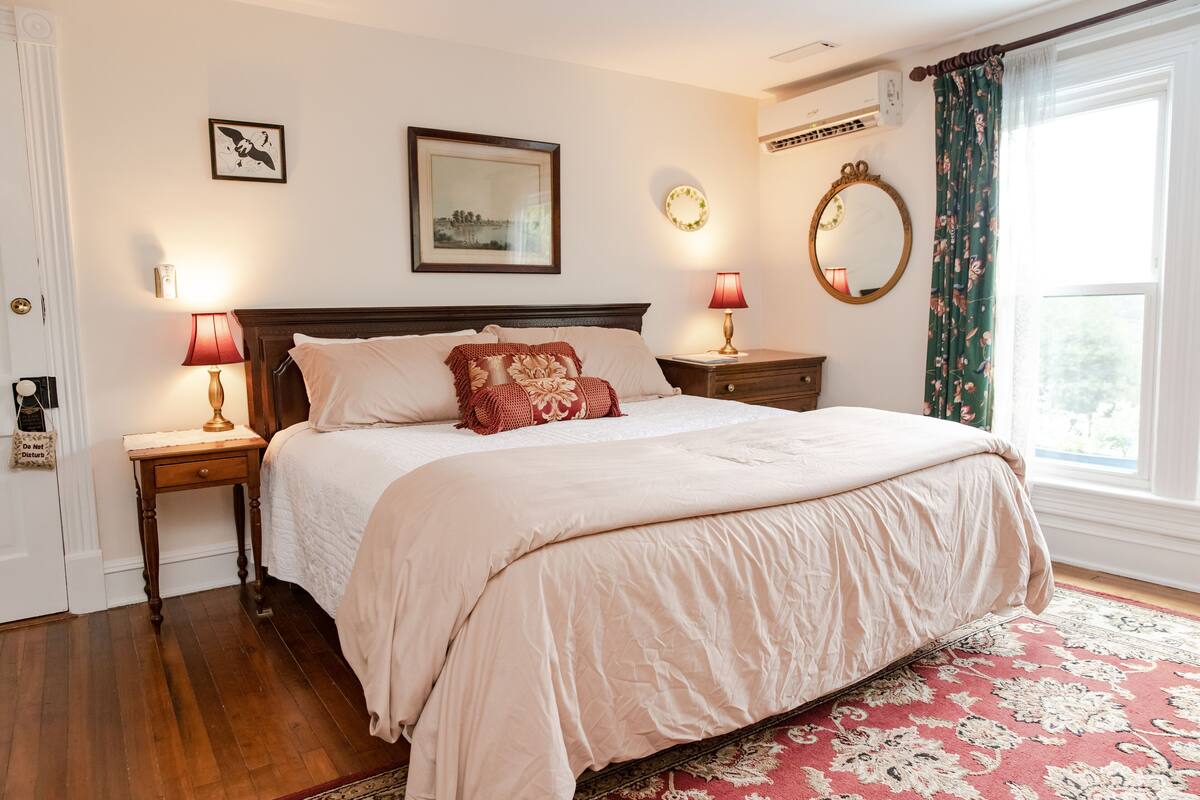 Une chambre à coucher bien aménagée, avec un grand lit, deux tables de chevet avec des lampes, un miroir fixé au mur et une fenêtre avec des rideaux.