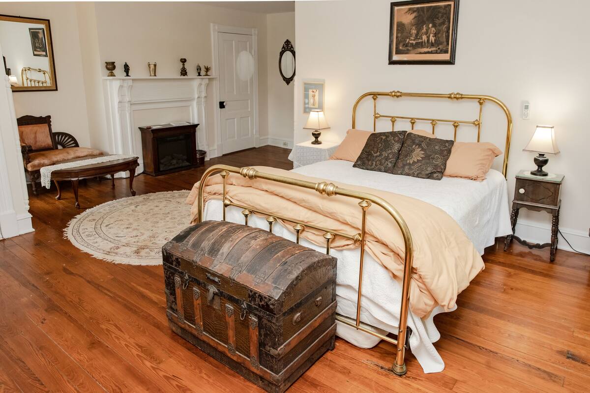 Une chambre avec un plancher en bois, un lit à cadre doré, une commode, une cheminée et un miroir au mur.