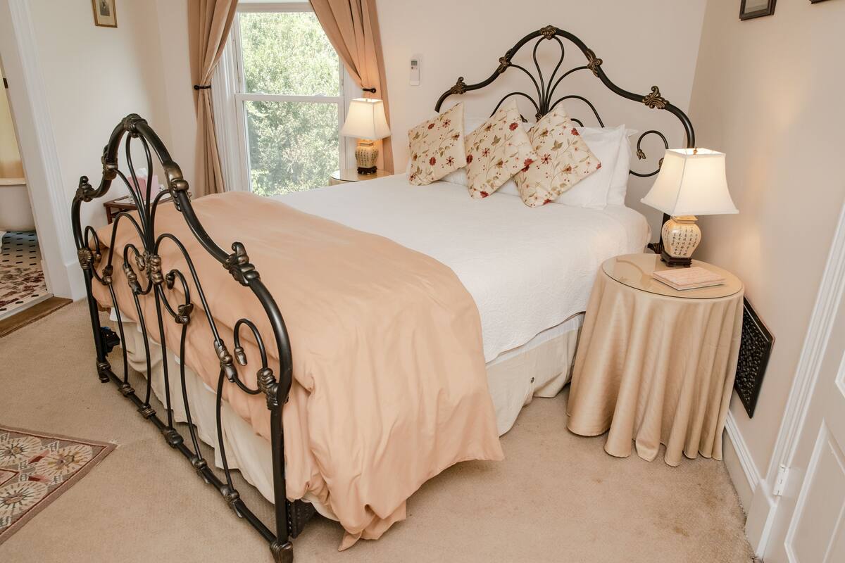 Un lit bien fait, agrémenté d’un ensemble d’oreillers à motifs floraux, une table de chevet avec une lampe et une vue sur l’extérieur par une fenêtre.