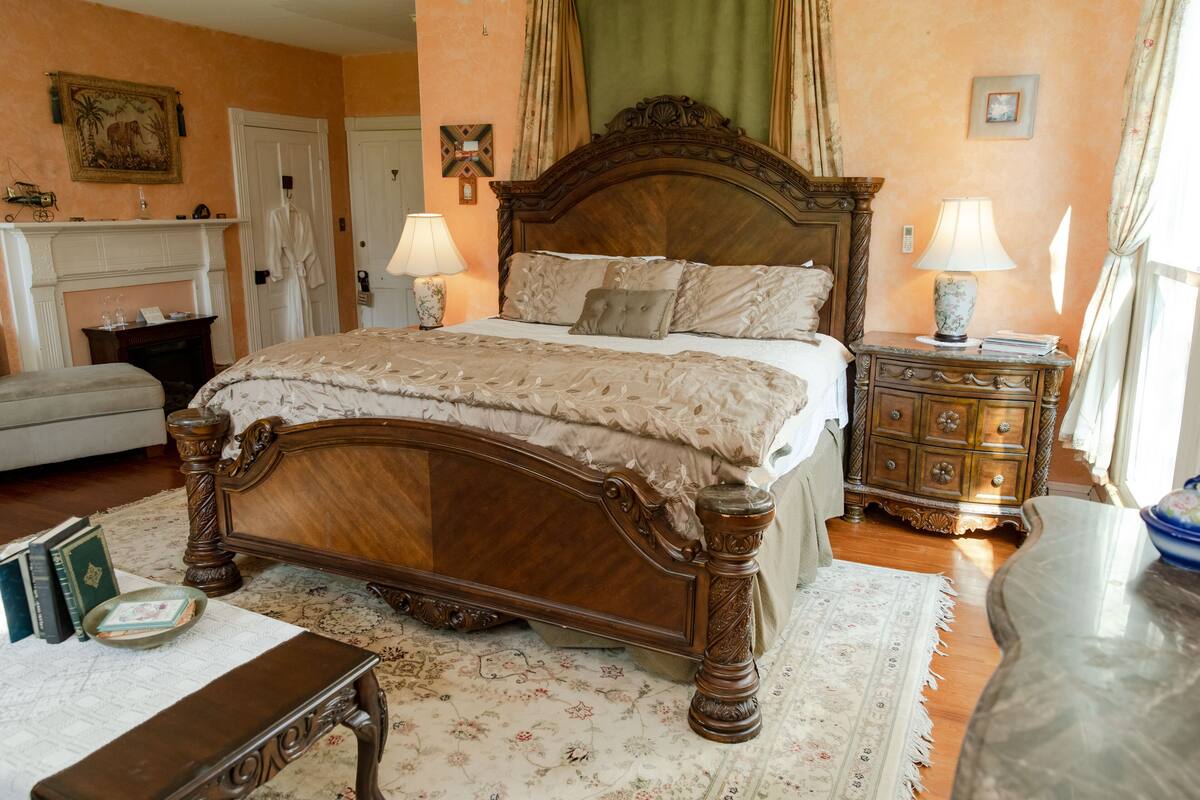 Une chambre à coucher de style traditionnel, avec un grand lit en bois, deux tables de chevet avec des lampes, une cheminée et une petite table avec des livres.