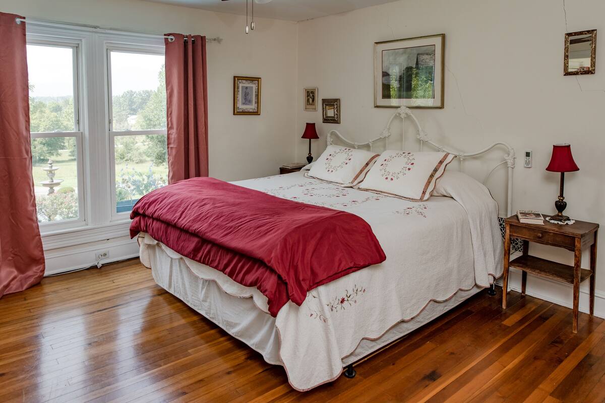 Une chambre avec un grand lit, un plancher en bois, une fenêtre avec des rideaux, une table de chevet avec une lampe et des tableaux encadrés accrochés au mur.