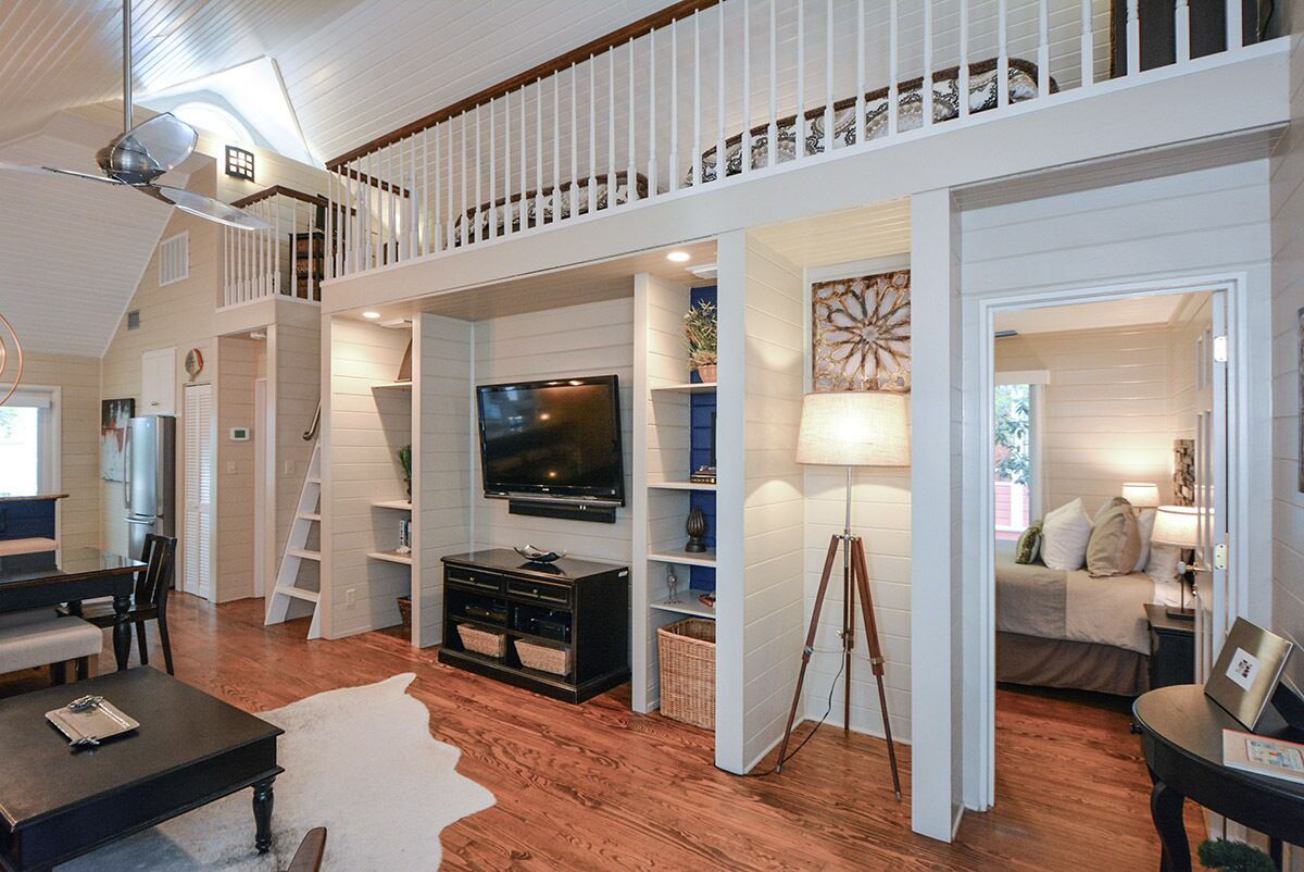 A spacious loft-style living area with a large TV, wooden floors, white walls, and a balcony.