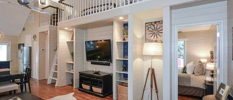 A spacious loft-style living area with a large TV, wooden floors, white walls, and a balcony.
