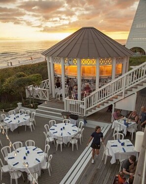 A gazebo with a roof, surrounded by outdoor tables and chairs, overlooking a beach.