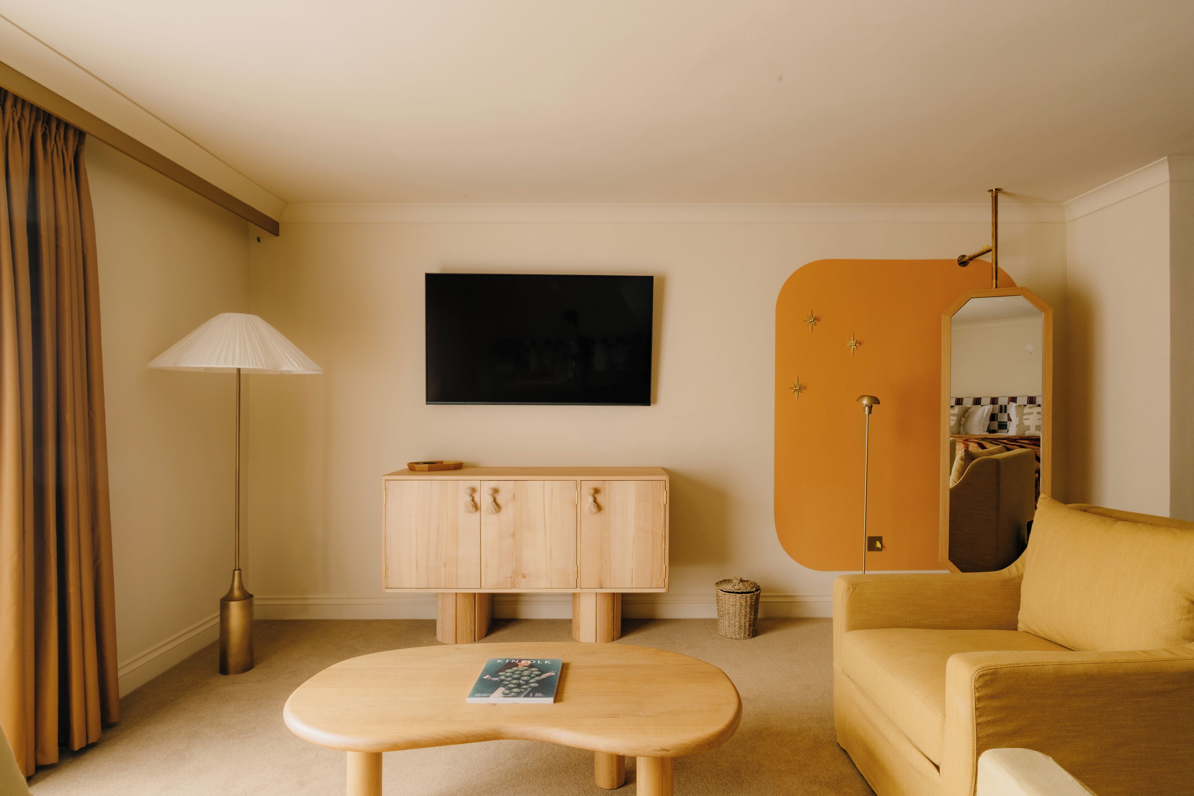 Habitación de hotel con un armario de madera, un sofá, una mesa con un libro, una lámpara de pie, un televisor montado en la pared y un espejo.