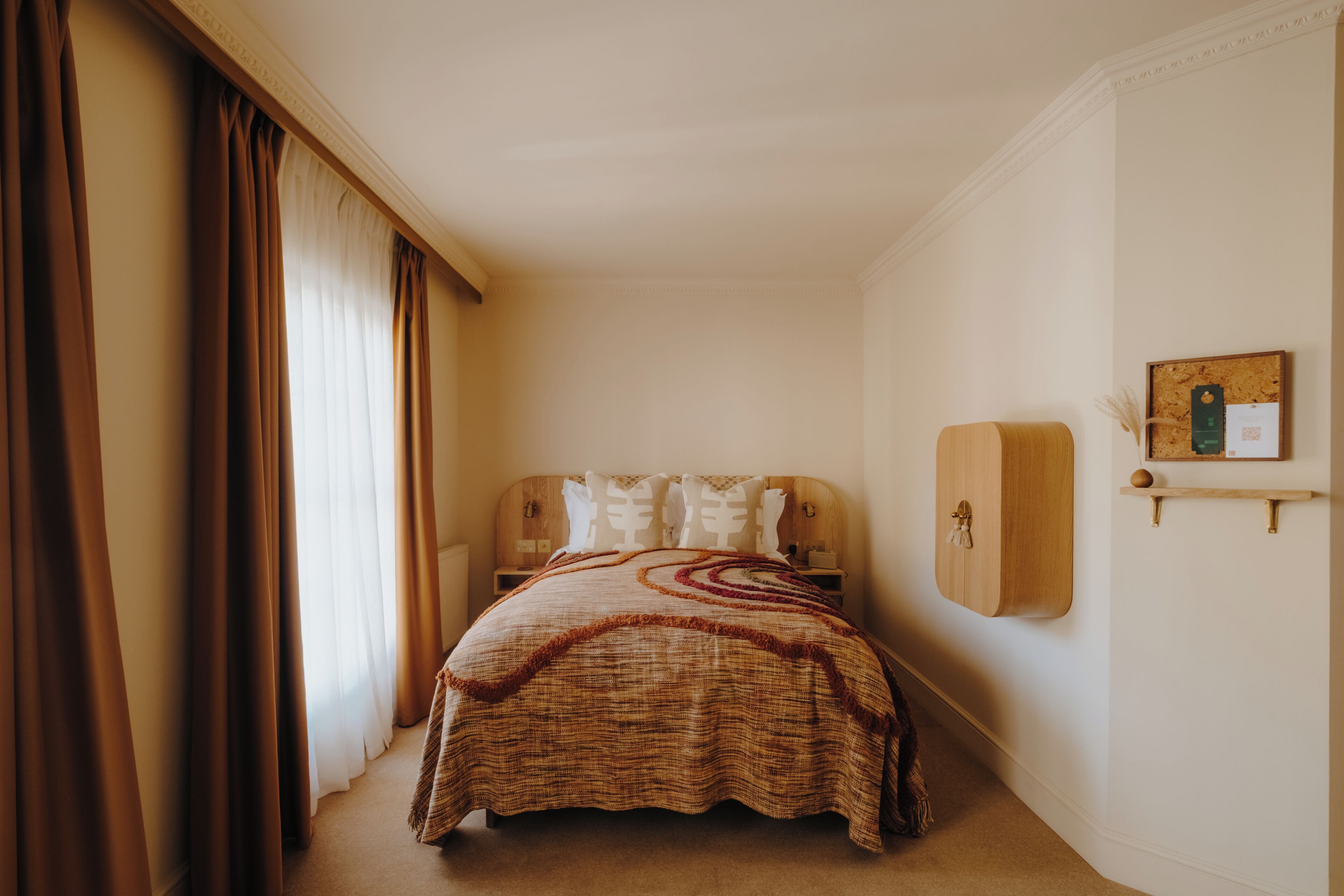 A bedroom with a bed, curtains, a nightstand, a wooden headboard, a wall-mounted shelf, and a corkboard.