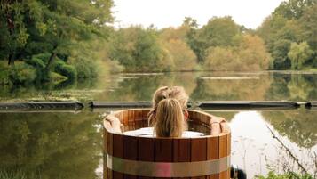 A wooden hot tub by a calm river with trees in the background.