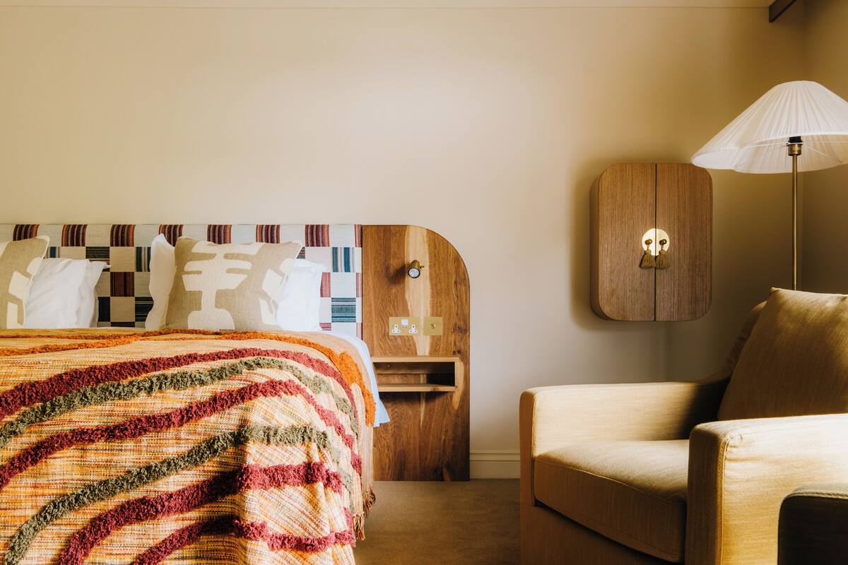 Ein Hotelzimmer mit einem Bett, einem Sessel, einem Nachttisch, einer Lampe und einem Holzkasten.