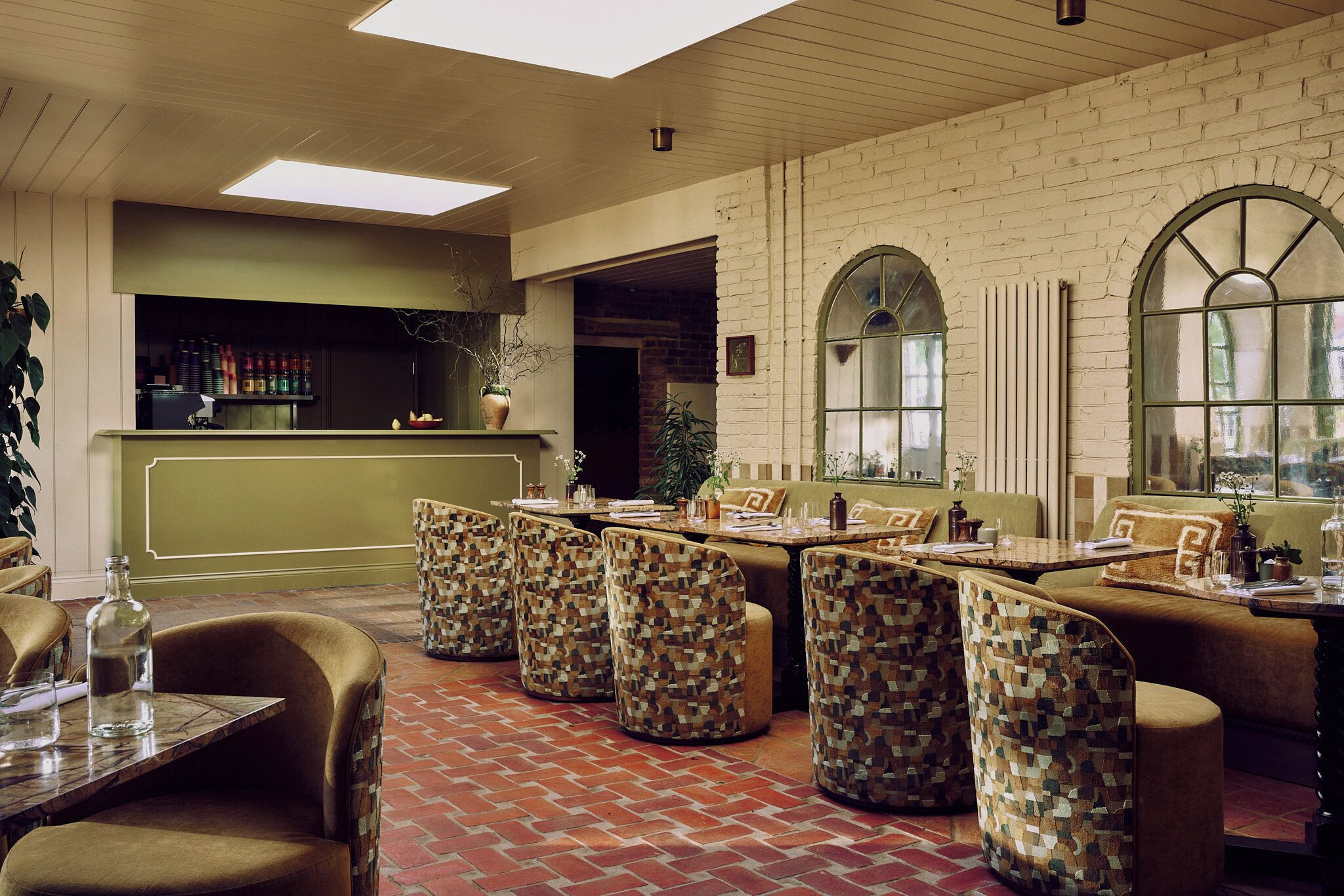 A restaurant with brick flooring, cushioned booths, a bar area, and large arched windows.