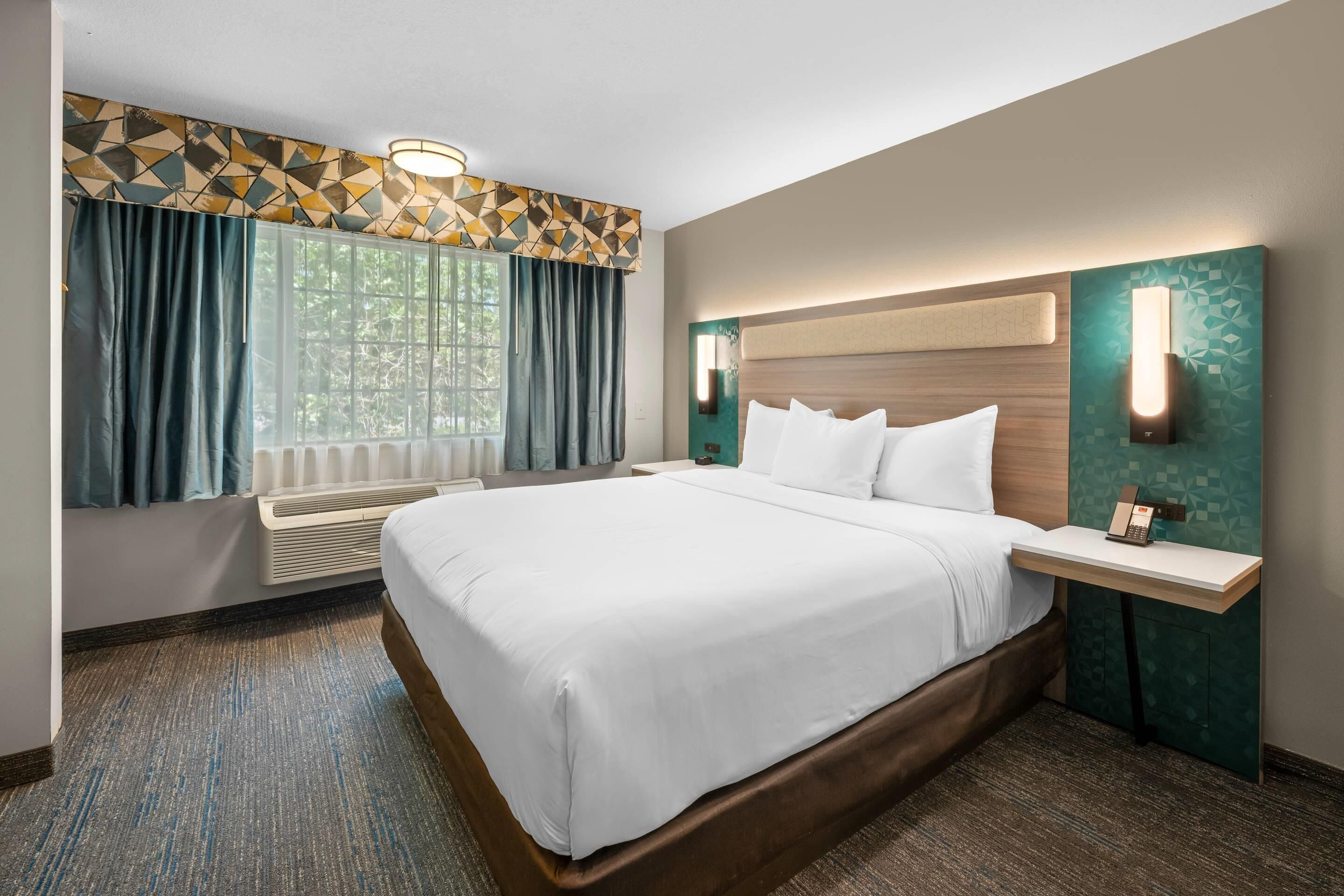 A hotel room with a large bed, a bedside table with a phone, a window with curtains, and a geometric-patterned wall.