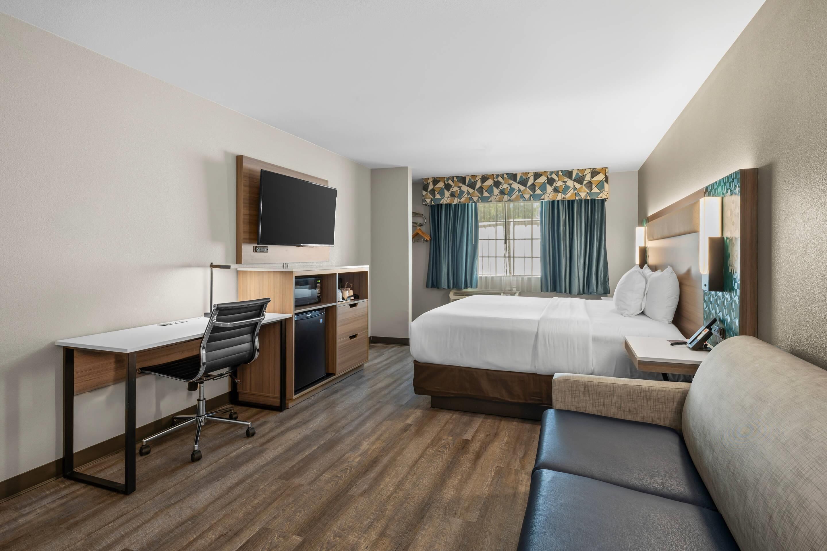 Une chambre d’hôtel avec un lit, un bureau, une chaise, une télévision, un canapé et une fenêtre avec des rideaux.
