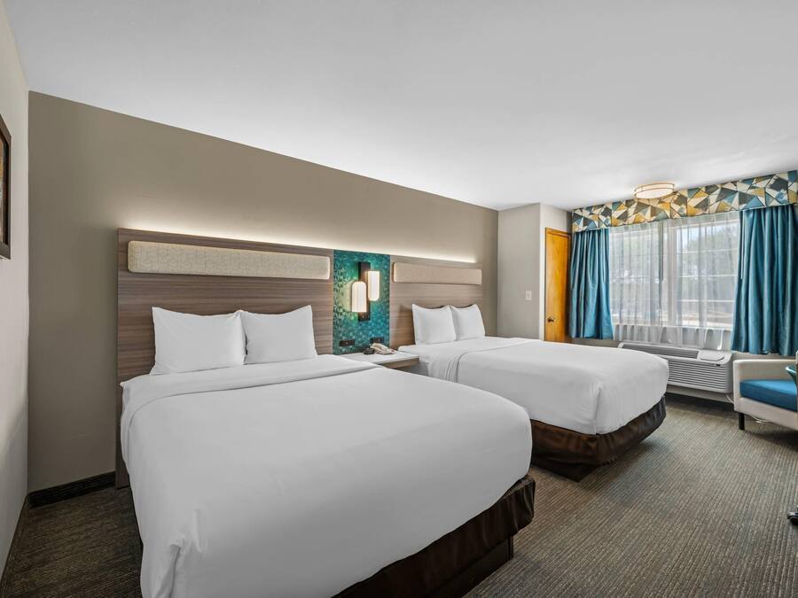 Ein Hotelzimmer mit zwei Betten, einem Schreibtisch, einem Stuhl, einem Fenster mit Vorhängen und einer Wandleuchte.