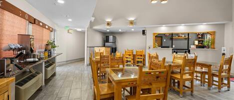 Un coin repas avec des tables et des chaises en bois, une cuisine à l’arrière-plan et un mur de briques sur le côté gauche.