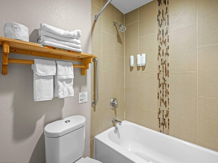 A bathroom with a toilet, a bathtub, a wooden shelf with towels, and a shower with a handrail.