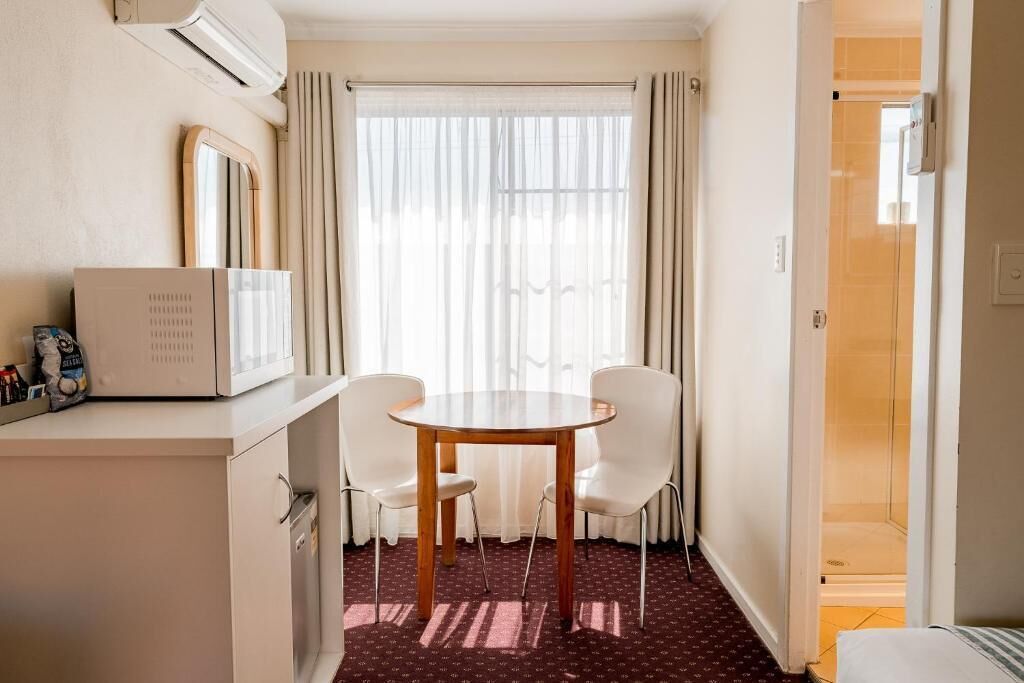 A hotel room with a dining table, two chairs, a microwave, a small cabinet, a window with curtains, and a shower area.