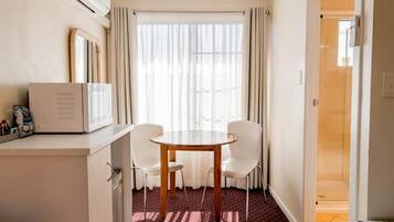 A hotel room with a dining table, two chairs, a microwave, a small cabinet, a window with curtains, and a shower area.