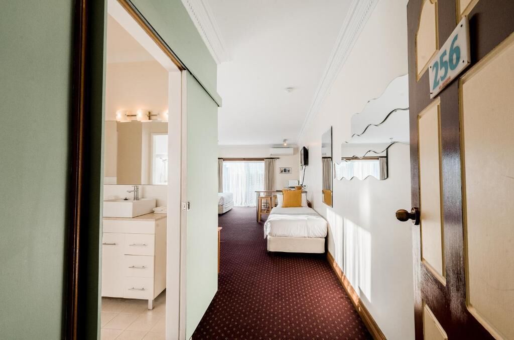A hotel room with a bed, a desk, a chair, a window, and a bathroom with a sink and mirror.