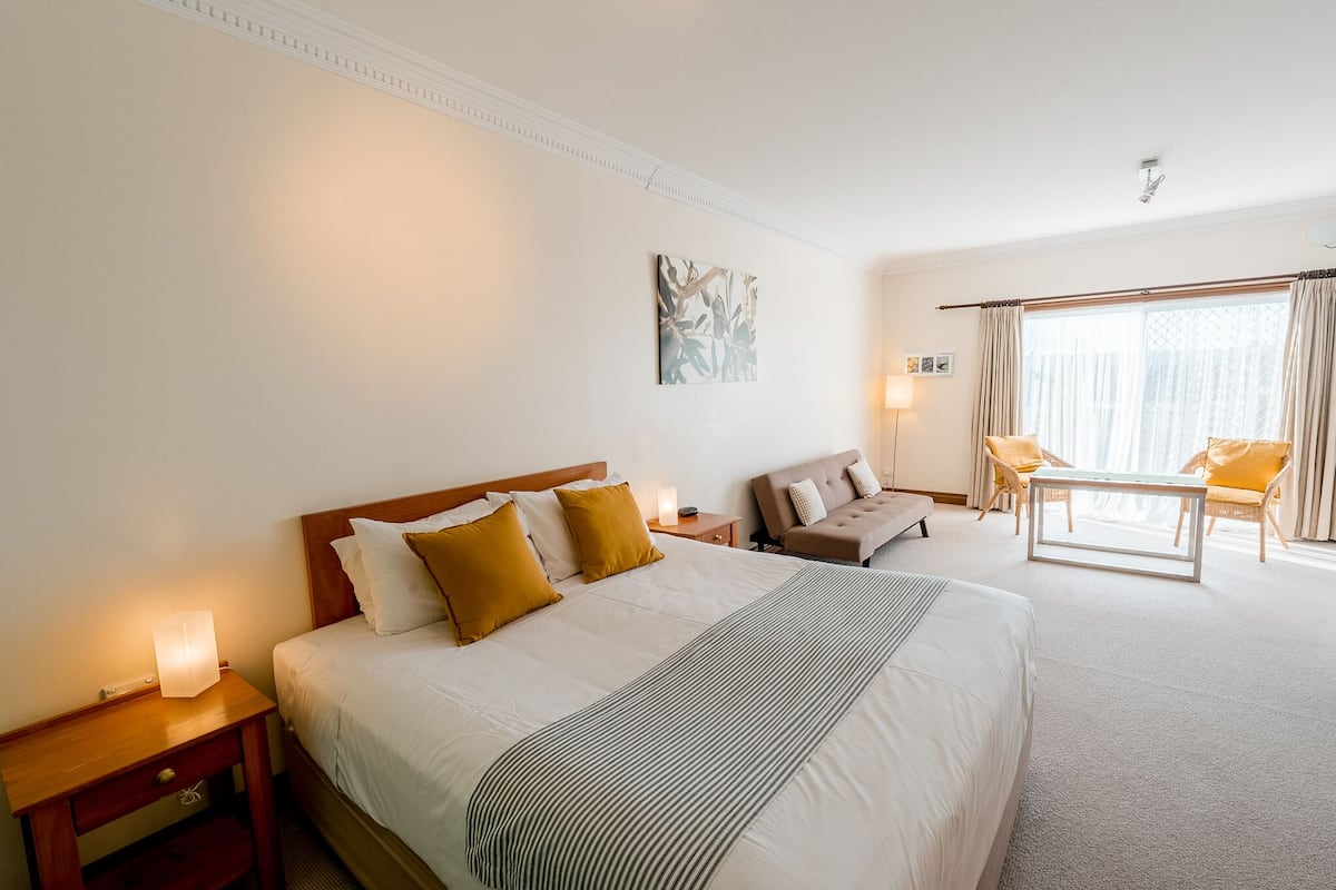 A hotel room with a large bed, a sofa, a small table, and a chair. There is a window with curtains, a nightstand with a lamp, and a wall-mounted artwork.
