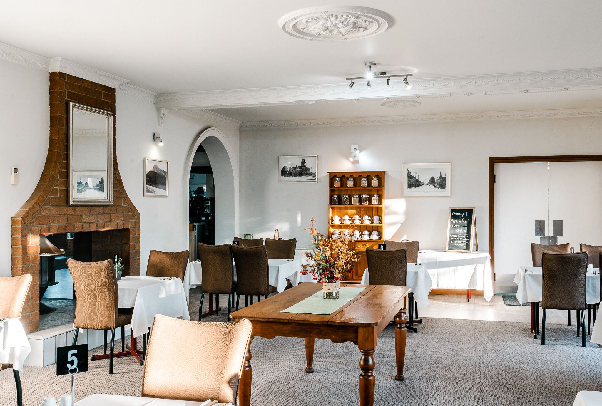 A dining area with a brick fireplace, white tables, brown chairs, and a decorative shelf with pottery.