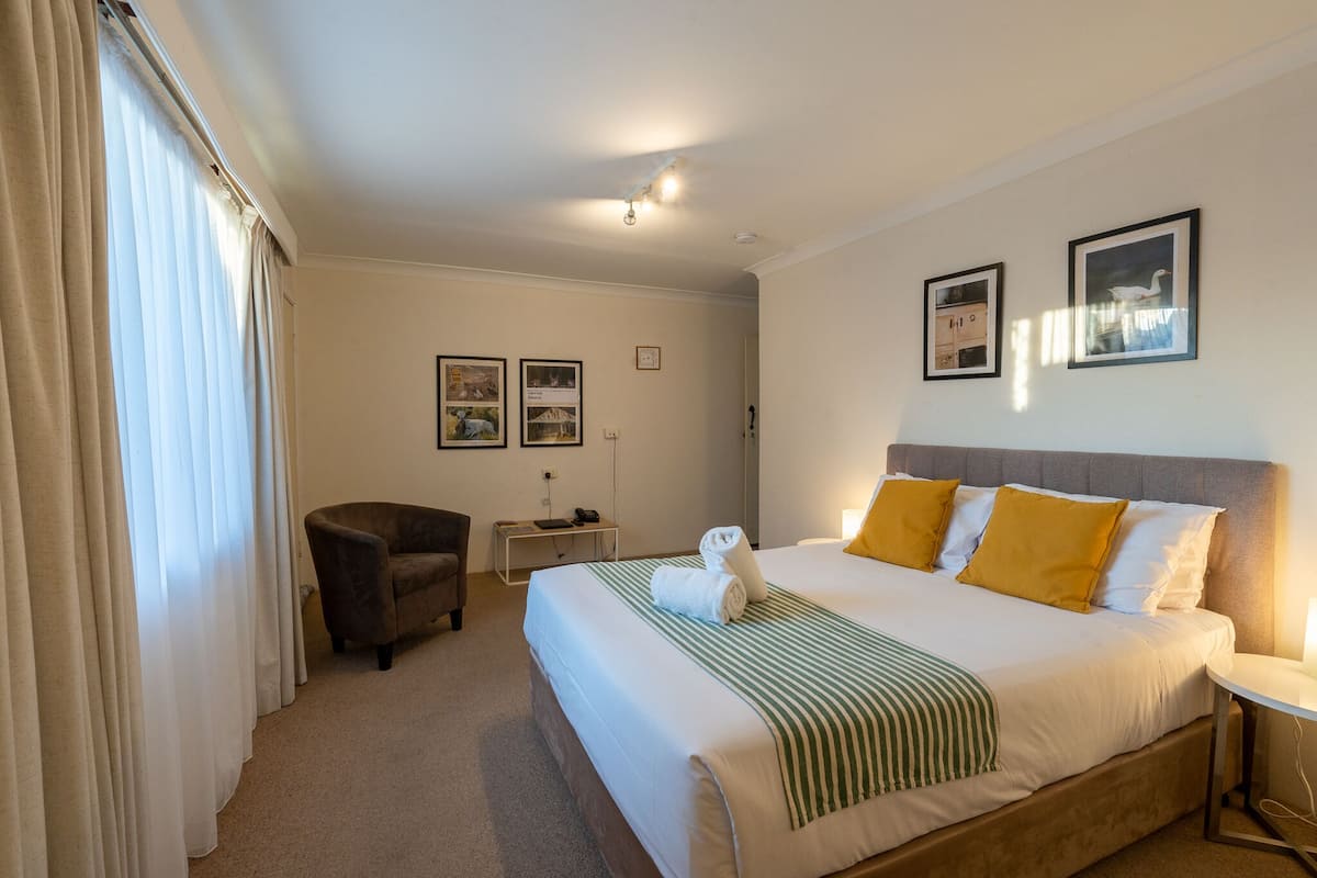 A hotel room with a large bed, a chair, a desk, and two framed pictures on the wall.