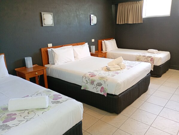 Habitación de hotel con dos camas individuales y una cama más grande. Cada cama tiene ropa de cama blanca y una colcha con estampado floral. Hay mesitas de noche de madera con lámparas y una pequeña mesa con una lámpara. La habitación tiene paredes oscuras y una ventana con cortinas.