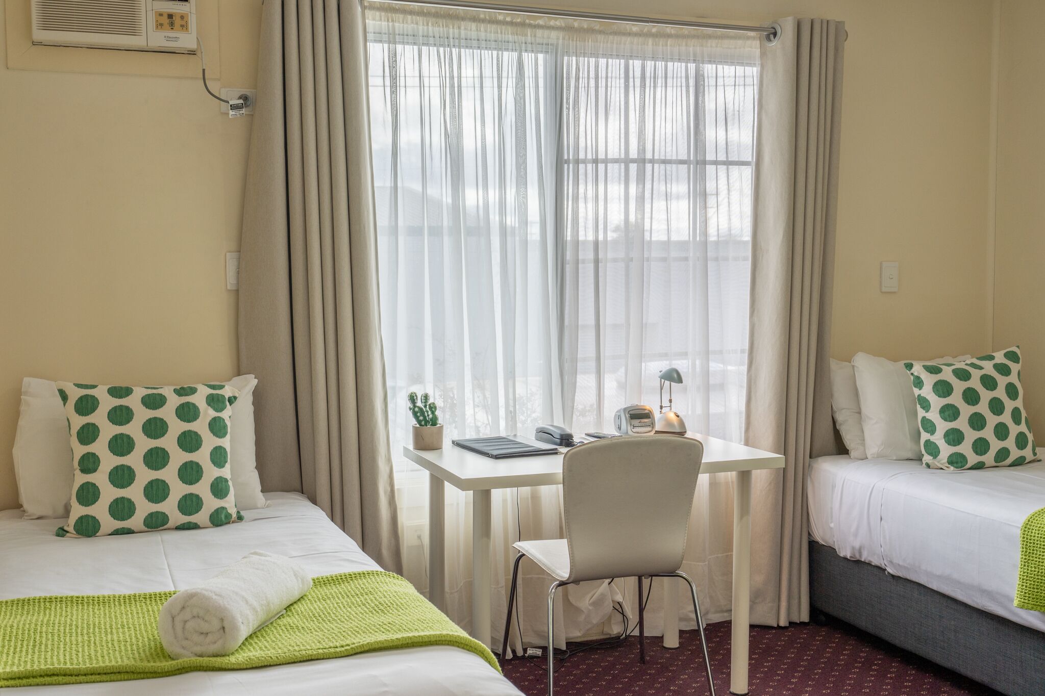 Et hotelv ærelse med to senge, et skrivebord, en stol, et lille bord med lampe og et vindue med let gardin.